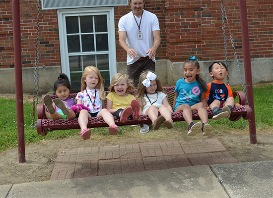 Six young children with hearing loss laugh as they sit on a swing together