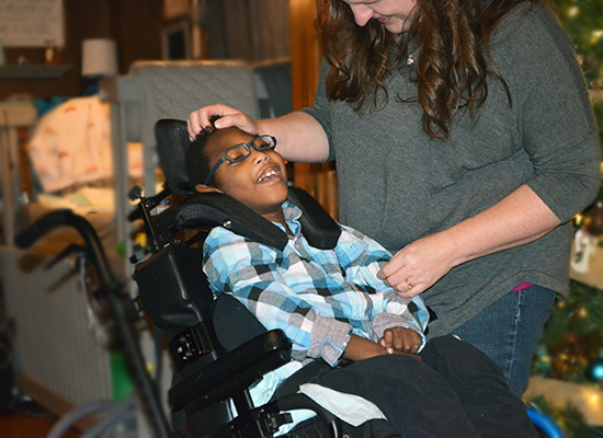 A boy with medical complexity sits in his wheelchair and smiles as his mother caresses his head