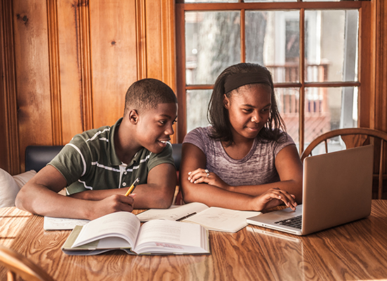 A brother and sister look at a laptop screen together while doing homework