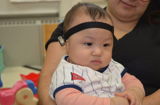 A toddler boy wearing a hearing aid device sits on a woman's lap