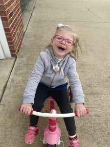 Home Care participant Willa sits on a pink tricycle and smiles up at the camera