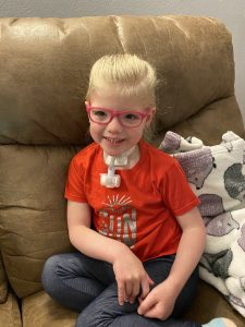 Home Care participant Willa sits on an armchair and smiles. She has pink glasses and a tracheostomy