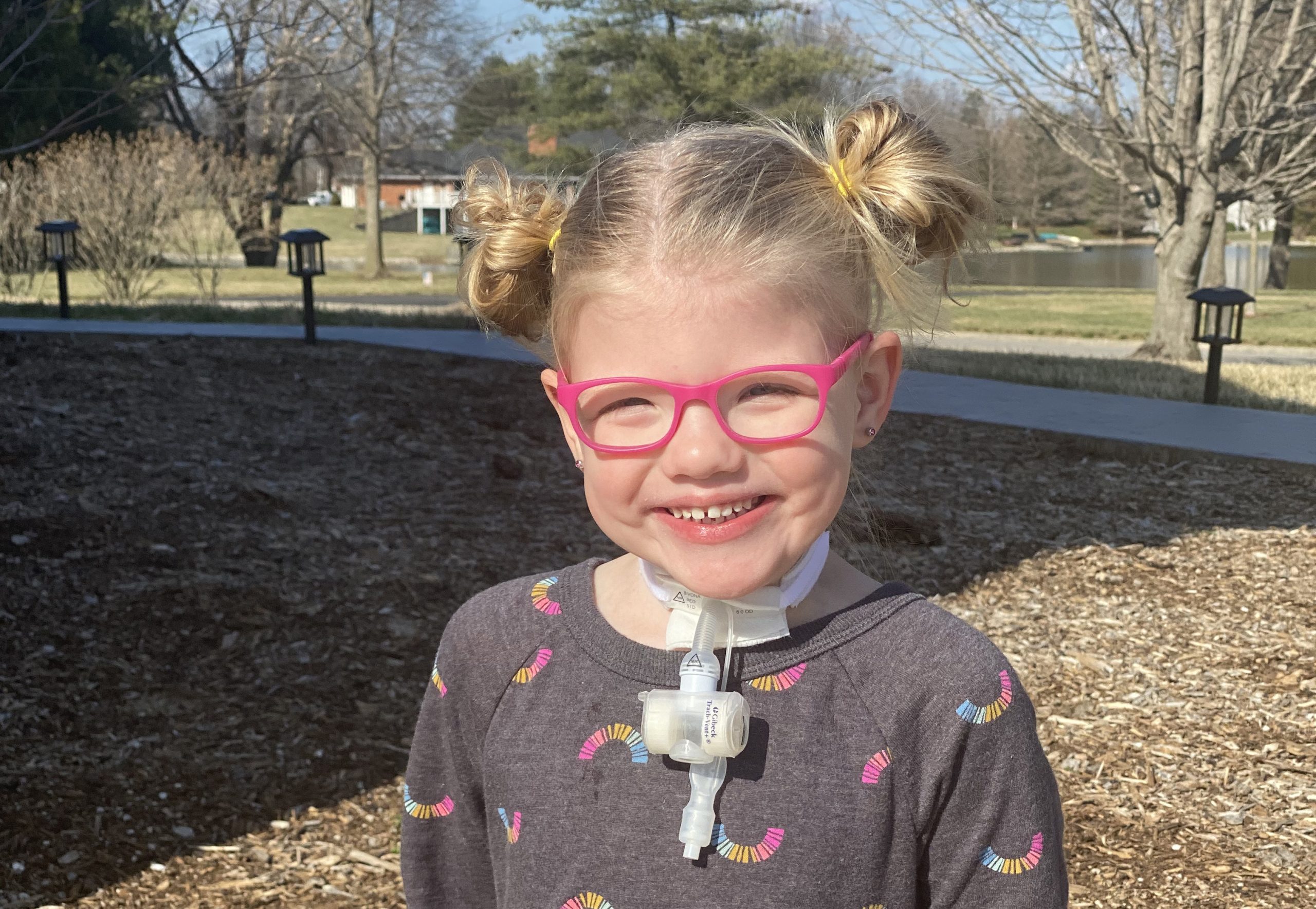 Home Care participant Willa sits outside with a big smile. She is wearing pink glasses and has a tracheostomy