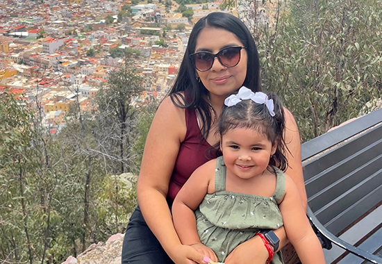 Current DSCC staff member and former participant Lisette Rios holds her young daughter in her lap as they sit on an outdoor ledge overlooking trees, hills and a city