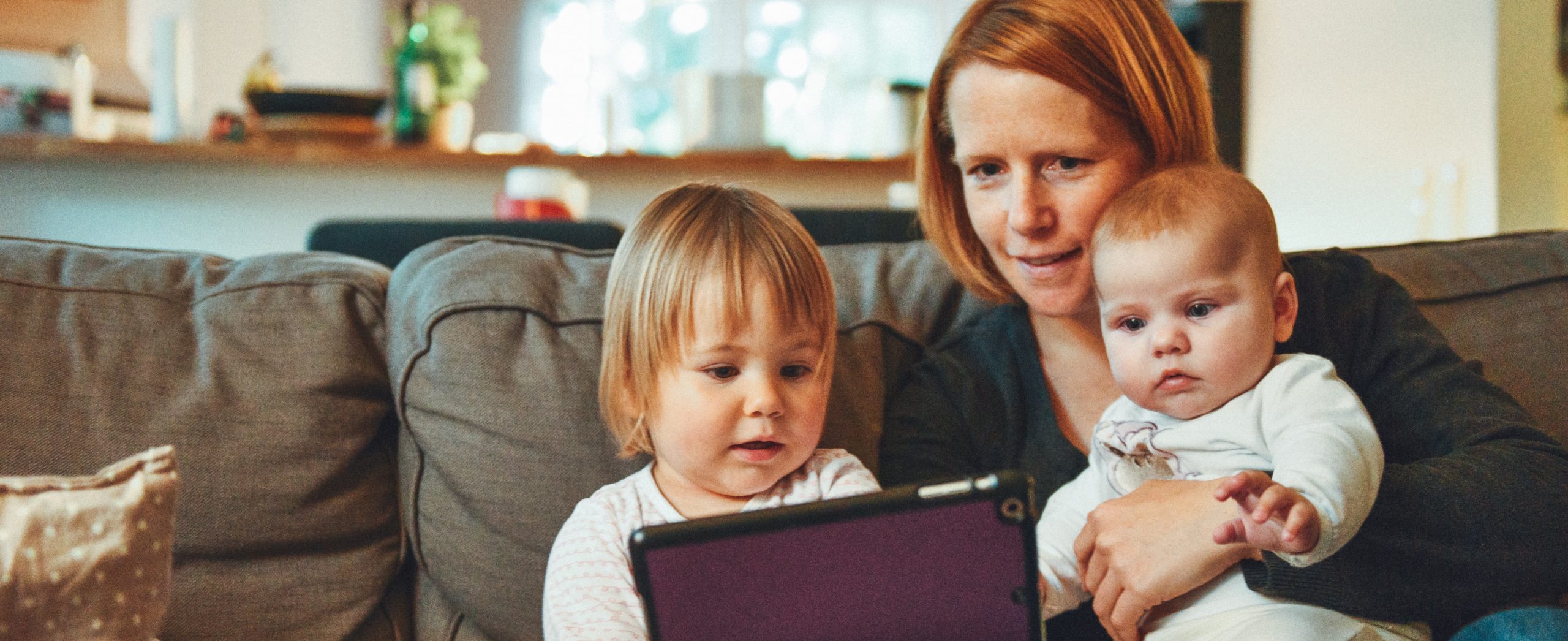 A mother with a baby on her lap and a toddler sitting beside her looks at a tablet screen
