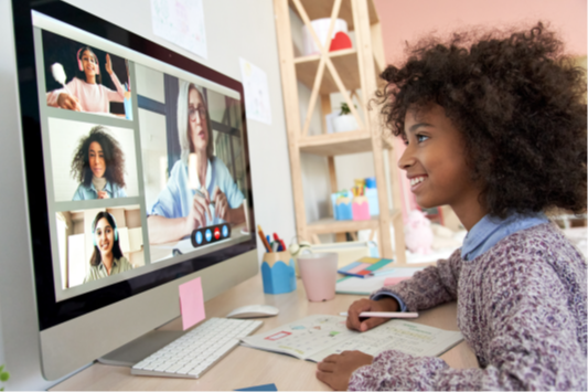 A dark-skinned teen girl sits at a desk and smiles at her large computer monitor as she participates in a virtual meeting