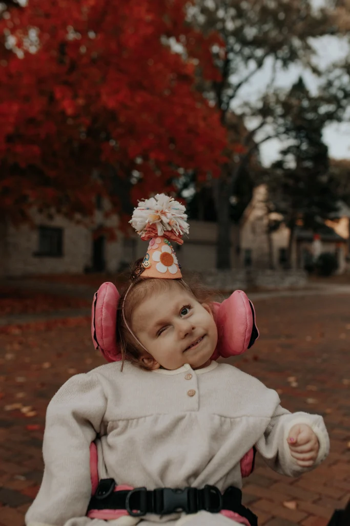 Eloise is wearing a light grey, long-sleeve fleece top and a cone-shipped birthday hat. The hat is made of light orange paper and that is decorated with a peace sign, flowers, a smiley face and a pom-pom on top. Her wheelchair has bright pink support pads that really pop in this picture.