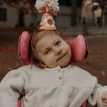 Eloise is wearing a light grey, long-sleeve fleece top and a cone-shipped birthday hat. The hat is made of light orange paper and that is decorated with a peace sign, flowers, a smiley face and a pom-pom on top. Her wheelchair has bright pink support pads that really pop in this picture.