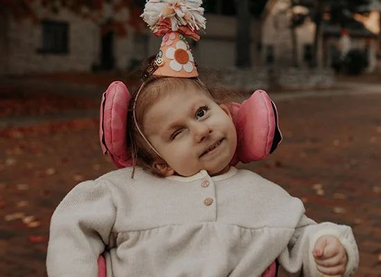 Eloise is wearing a light grey, long-sleeve fleece top and a cone-shipped birthday hat. The hat is made of light orange paper and that is decorated with a peace sign, flowers, a smiley face and a pom-pom on top. Her wheelchair has bright pink support pads that really pop in this picture.