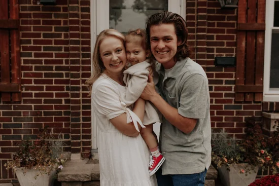 Rachel and Zach Singelton hold their 3-year-old daughter Eloise closely between them while standing outdoors on a front porch. All three have big smiles.