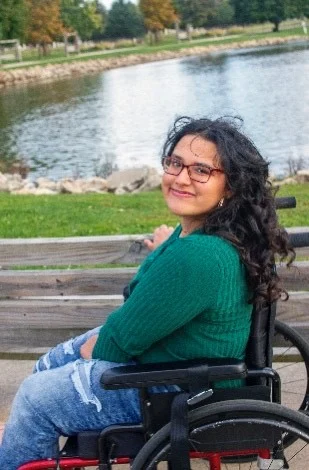 DSCC participant Petra Petty, 17, sits in her wheelchair and smiles at the camera while sitting outdoors near a pond