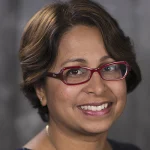 Dr. Shubhra Mukherjee has a beautiful smile, brown eyes and short, dark brown hair that frames her face. She is wearing glasses. The frames are dark red and are shaped like an oval that has been slightly squared on one end. She is wearing a navy colored, sleeveless top that has a honeycomb pattern woven into the fabric