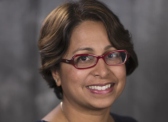 Dr. Shubhra Mukherjee has a beautiful smile, brown eyes and short, dark brown hair that frames her face. She is wearing glasses. The frames are dark red and are shaped like an oval that has been slightly squared on one end. She is wearing a navy colored, sleeveless top that has a honeycomb pattern woven into the fabric