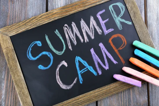 The text, "Summer Camp," written with chalk on chalkboard next to chalk sticks of different bright colors