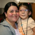 Mom Maria holds her daughter Dalilah, a young girl with hearing loss who wears glasses and hearing aids. They are both smiling at the camera