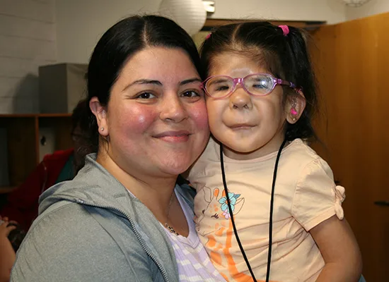 Mom Maria holds her daughter Dalilah, a young girl with hearing loss who wears glasses and hearing aids. They are both smiling at the camera