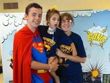 DSCC participant Isabella's older brother Braden wears a Superman costume and holds Isabella in his arms next to Isabella's nurse, Toni. A superhero comic backdrop is behind them.