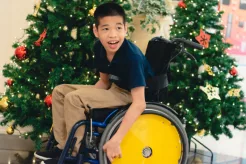An older, dark-haired boy in a wheelchair smiles at the Christmas trees and holiday decorations that surround him
