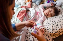 A mother administers food through her young daughter's gastrostomy tube while the little girl lies on her bed