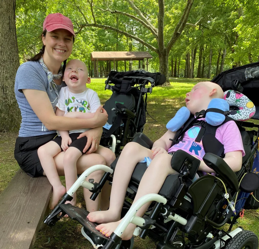 Erica Stearns sits outside on a picnic bench and holds her son Caratacus in her lap while sitting next to her daughter Margot, who is in a medical stroller