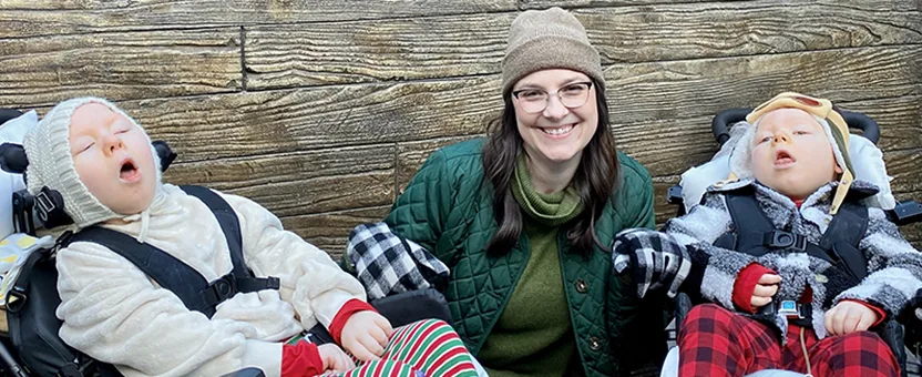 Erica Stearns kneels between her two children, Margot and Caratacus, as they sit in medical stroller outdoors. All three are wearing winter hats and coats.