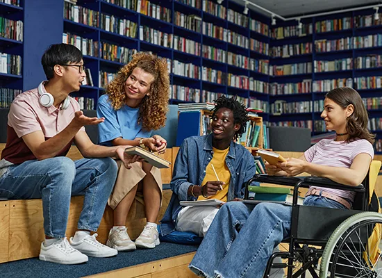A diverse group of four students, including a young woman in a wheelchair, gathered in a college library and enjoying discussion