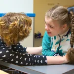 A nurse sits face-to-face with and extends her arms around a girl with complex medical needs