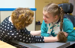A nurse sits face-to-face with and extends her arms around a girl with complex medical needs