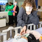 A woman in a mask checking the tracheostomy tube of an infant medical training mannequin in a simulation room setting