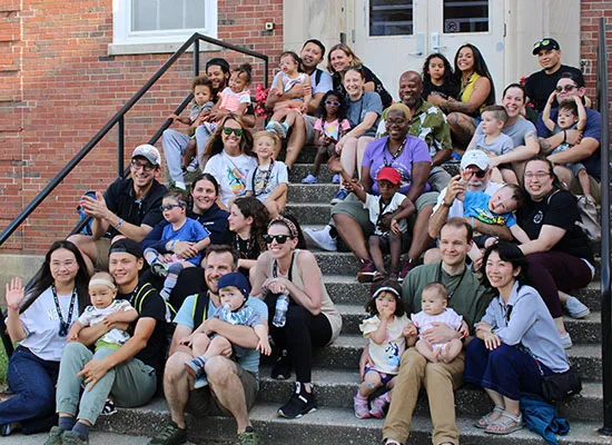 A group of 12 families who attended the 2024 Institute for Parents of Preschool Children Who Are Deaf or Hard of Hearing pose together on the steps of the dorms on the Illinois School for the Deaf campus.