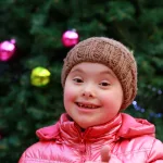 A young girl with Down syndrome wearing a winter coat and hat standing outdoors in front of a Christmas tree