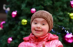 A young girl with Down syndrome wearing a winter coat and hat standing outdoors in front of a Christmas tree