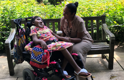 Ekiko "Yesenia" Aiken sits on a park bench outdoors and smiles at her daughter, Davina, who is in a wheelchair and facing her while holding her hand