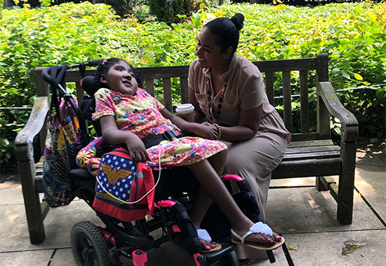 Ekiko "Yesenia" Aiken sits on a park bench outdoors and smiles at her daughter, Davina, who is in a wheelchair and facing her while holding her hand