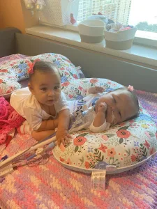 Ava and Olivia Vasquez are infants, lying together on colorful baby pillows. Ava has a tracheostomy tube. 