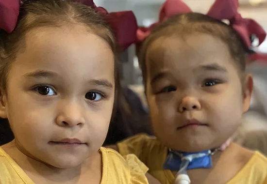 Twin toddlers Olivia and Ava Vasquez look at the camera. Ava is medically complex and has a tracheostomy tube.