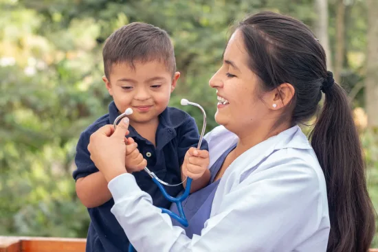 A woman wearing a white jacket and scrubs holds a young boy with disabilities in her arms as they both hold a stethoscope and laugh together