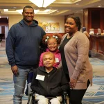 A family of four smiles together in the middle of the hotel lobby. The parents are on either side of their children. Their teen son is in front, he uses a wheelchair. His younger sister stands behind him.