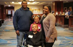 A family of four smiles together in the middle of the hotel lobby. The parents are on either side of their children. Their teen son is in front, he uses a wheelchair. His younger sister stands behind him.