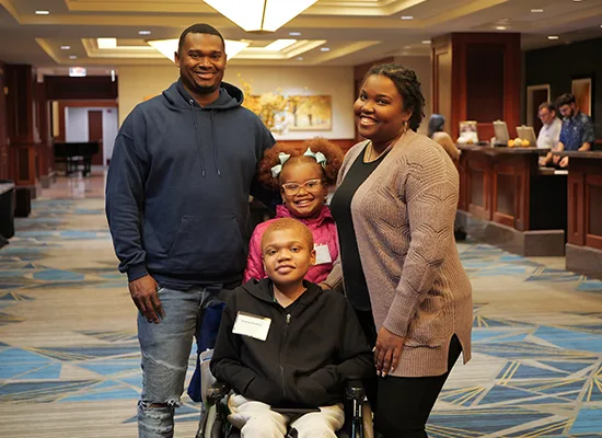 A family of four smiles together in the middle of the hotel lobby. The parents are on either side of their children. Their teen son is in front, he uses a wheelchair. His younger sister stands behind him.