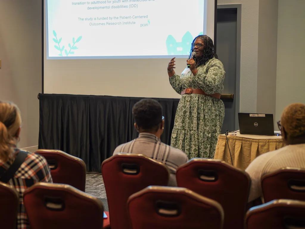 A DSCC team member gives a presentation to conference attendees. She holds a microphone while standing in front of a presentation slide. Conference attendees sit in the first and second rows.