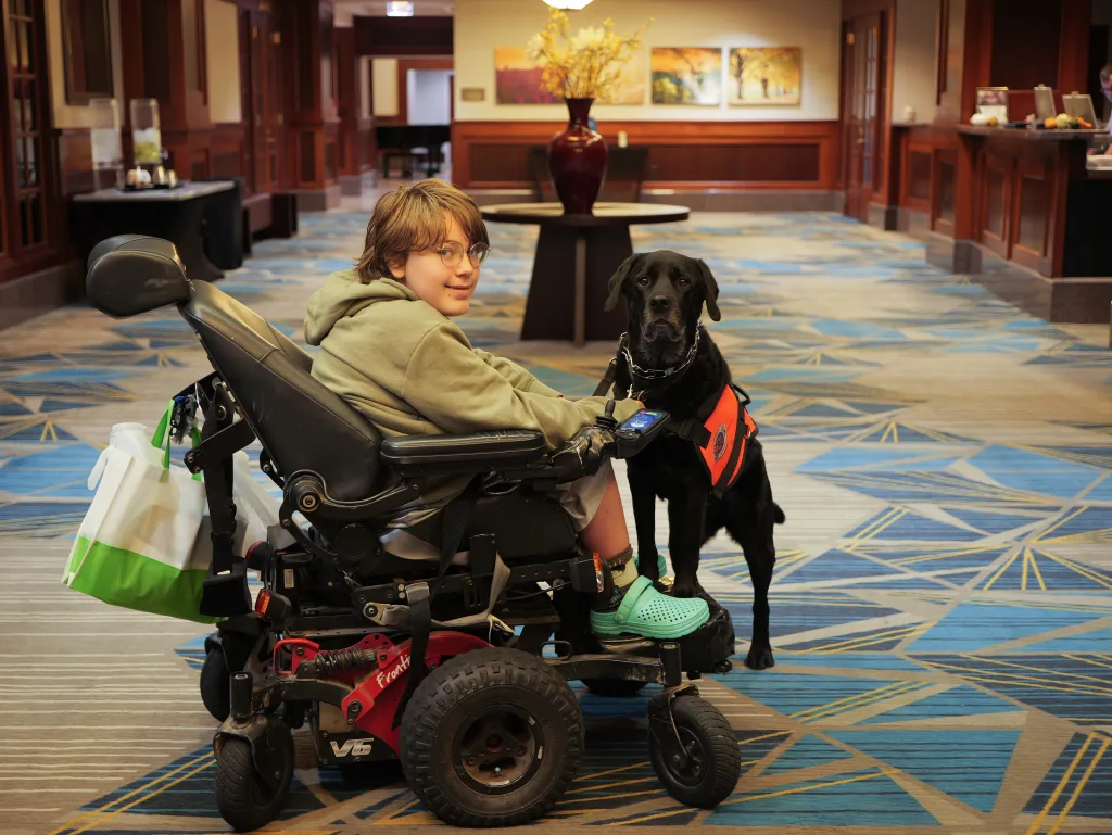 A conference attendee smiles in the middle of the hotel lobby. He uses an electric-powered wheelchair. His service dog is next to him.