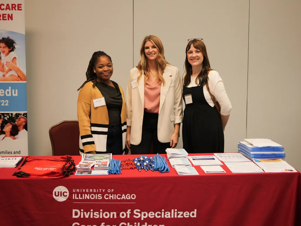 Three DSCC team members stand behind DSCC's table at the 2024 Transition Conference. The table has handouts, backpacks, pens, and other items to share with attendees. A DSCC banner stands upright behind them. It has photos of families and information about DSCC.  