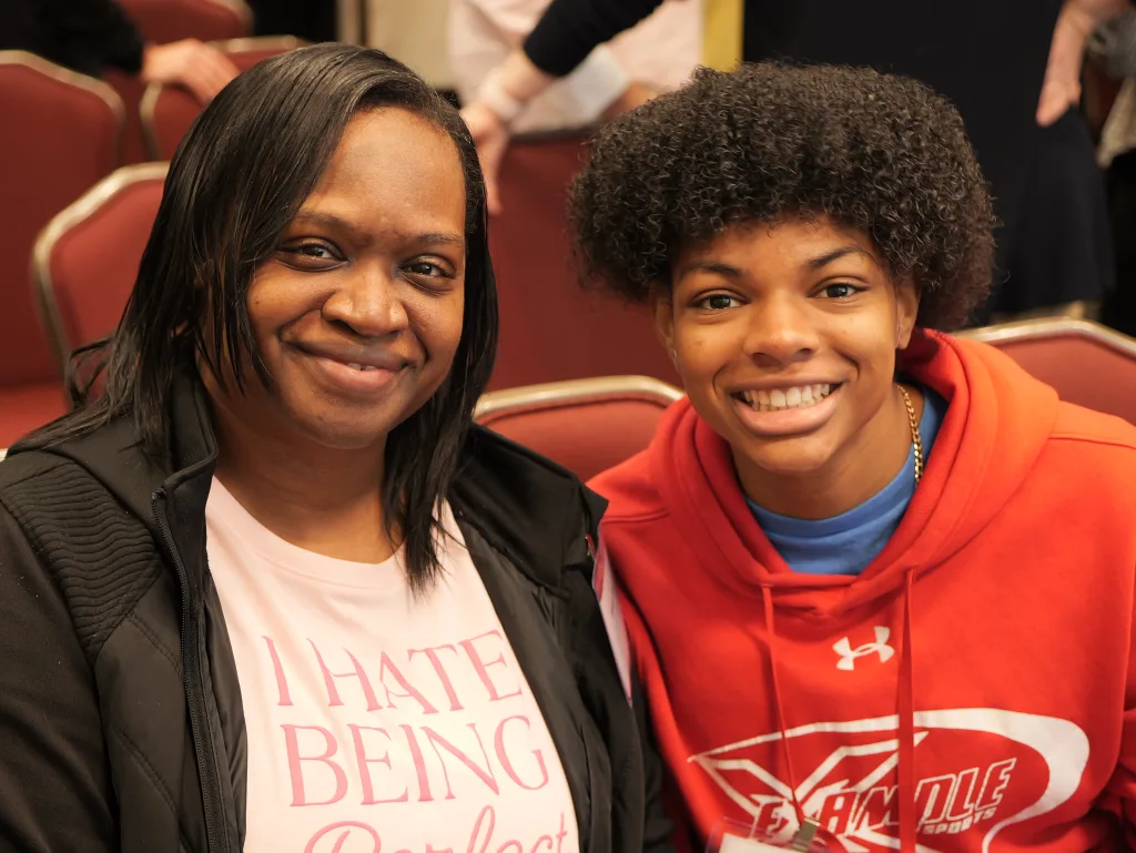 A mother and daughter sit next to each other during a meetup for DSCC participants. People stand around the chairs in the rows behind them. 