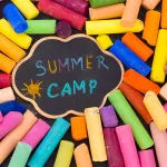 Colorful pieces of chalk surround a small cloud-shaped chalkboard. Blue, yellow, purple and turquoise chalk was used to draw a sun and write "Summer Camp" on the chalkboard.