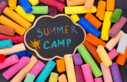 Colorful pieces of chalk surround a small cloud-shaped chalkboard. Blue, yellow, purple and turquoise chalk was used to draw a sun and write "Summer Camp" on the chalkboard.