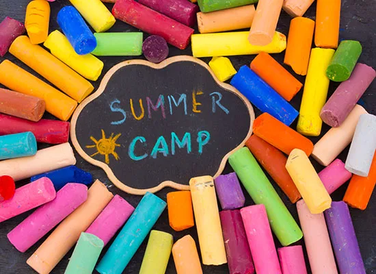 Colorful pieces of chalk surround a small cloud-shaped chalkboard. Blue, yellow, purple and turquoise chalk was used to draw a sun and write "Summer Camp" on the chalkboard.