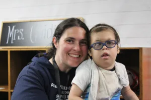 A mom hugs her four-year-old son outside of his classroom. He wears glasses and has hearing aids.