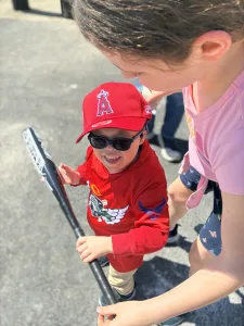 A smiling four-year-old wears sunglasses and a baseball uniform. He holds a baseball bat. His older sister learns over him, holding onto the end of the bat.