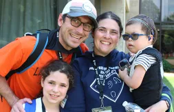 A family of four smiles together in front of the Illinois School for the Deaf. The mom and dad are in the center of the photo. The mom holds her four-year-old son. He wears glasses and has hearing aids. The dad also has glasses. He puts his hand on the shoulder of his seven-year-old daughter.